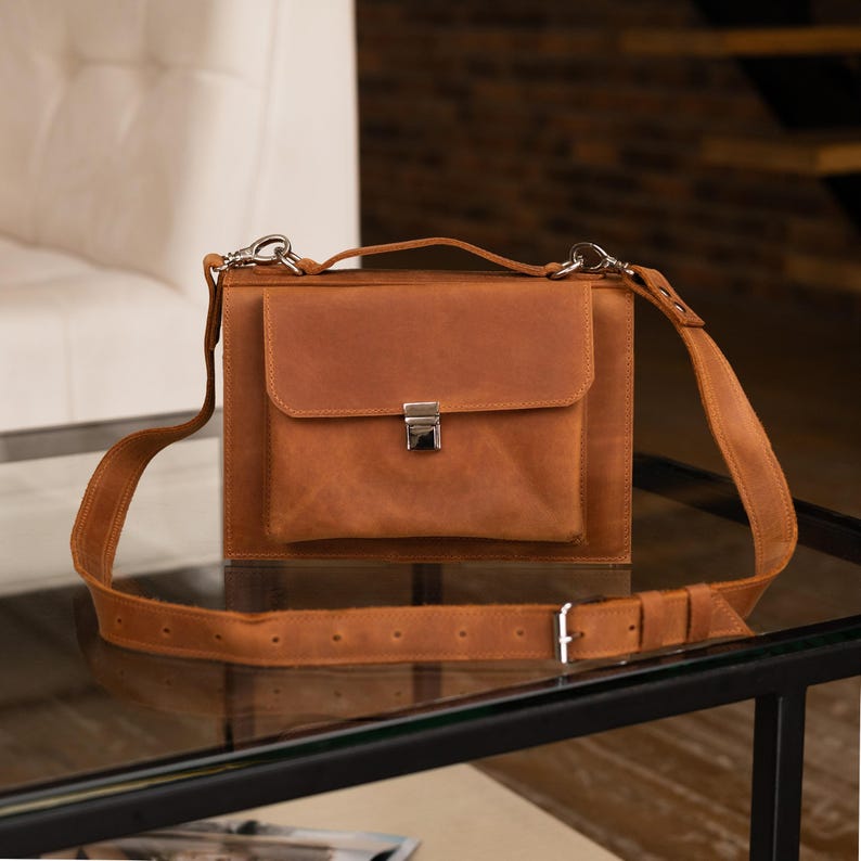 K&ouml;nnte beinhalten: Eine braune Ledertasche mit einer Klappentasche und einem silberfarbenen Verschluss. Die Tasche hat einen langen, verstellbaren Schulterriemen und einen oberen Griff. Die Tasche steht auf einer Glasoberfl&auml;che.