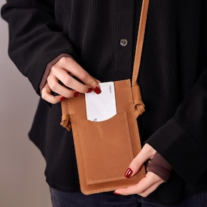 May include: A tan leather crossbody phone bag with a front pocket. A white card is being inserted into the bag. The bag has a long strap and is worn over a black jacket.