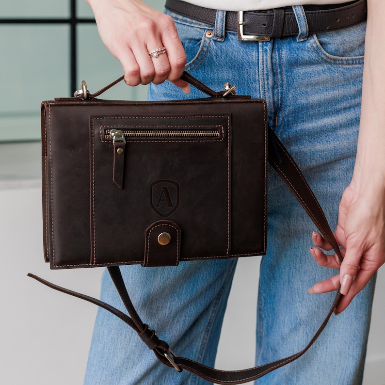 May include: A dark brown leather satchel with a top handle and a detachable shoulder strap. The satchel features a front zippered pocket, a flap closure with a snap, and an embossed letter "A" on the front. The bag is held by a person wearing blue jeans.