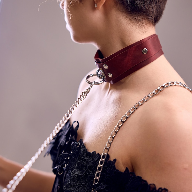 May include: A burgundy leather collar with silver-toned hardware and a matching chain. The collar is wide and secured with rivets. The chain is attached to a large metal ring on the collar.
