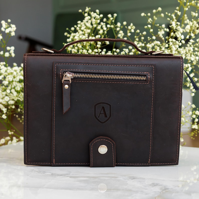 May include: Dark brown leather briefcase with a top handle and a front pocket with a zipper. The briefcase has a flap closure with a silver button and a debossed letter "A" on the front.