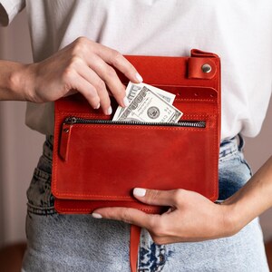Może przedstawiać: Czerwony sk&oacute;rzany portfel z zapięciem na zamek błyskawiczny i wieloma przegr&oacute;dkami. Ręka wkłada banknot 100 dolar&oacute;w do kieszeni zapinanej na zamek. Portfel jest prostokątny i ma zapięcie na klapę z zatrzaskiem.