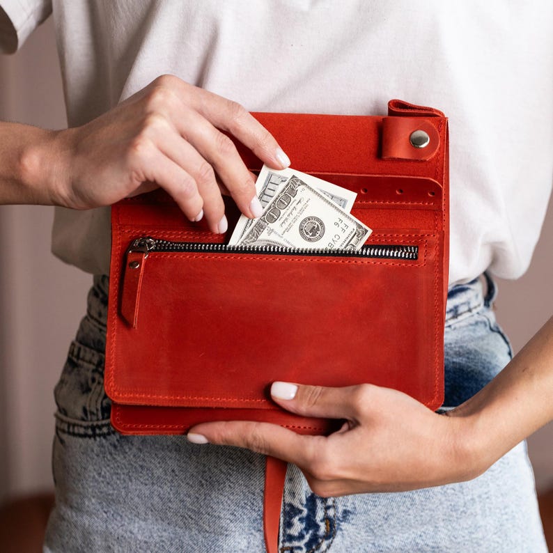 Może przedstawiać: Czerwony sk&oacute;rzany portfel z zapięciem na zamek błyskawiczny i wieloma przegr&oacute;dkami. Ręka wkłada banknot 100 dolar&oacute;w do kieszeni zapinanej na zamek. Portfel jest prostokątny i ma zapięcie na klapę z zatrzaskiem.