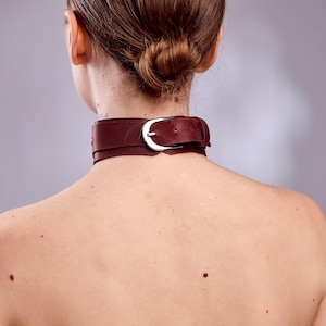 May include: A burgundy leather choker with a silver-colored buckle. The choker is worn around the neck. The image is taken from the back, showing the nape of the neck and the back of the head.