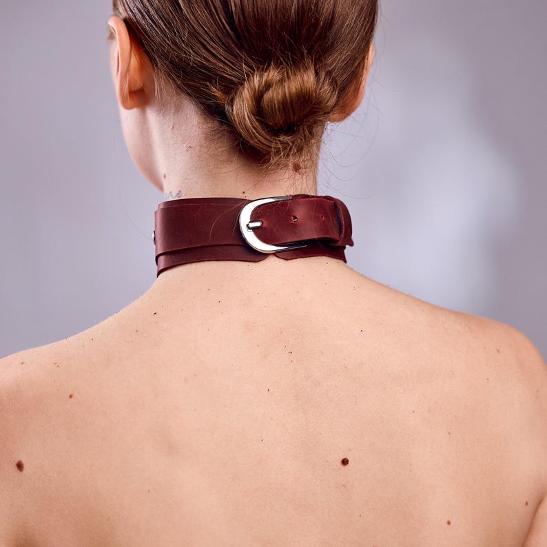 May include: A burgundy leather choker with a silver-colored buckle. The choker is worn around the neck. The image is taken from the back, showing the nape of the neck and the back of the head.