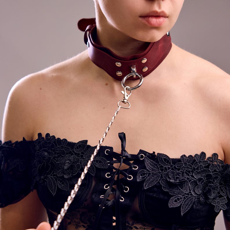 May include: A burgundy leather collar with silver-tone hardware and a matching leash. The collar is secured with a buckle and rivets. The black lace top is visible in the image.