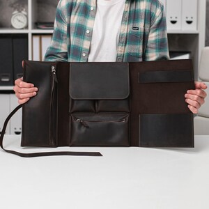 May include: A dark brown leather portfolio with multiple pockets and a zipper closure. The open portfolio displays a variety of compartments, including a large pocket, smaller pockets, and a pen holder. The leather has a smooth texture and a rich, dark color.