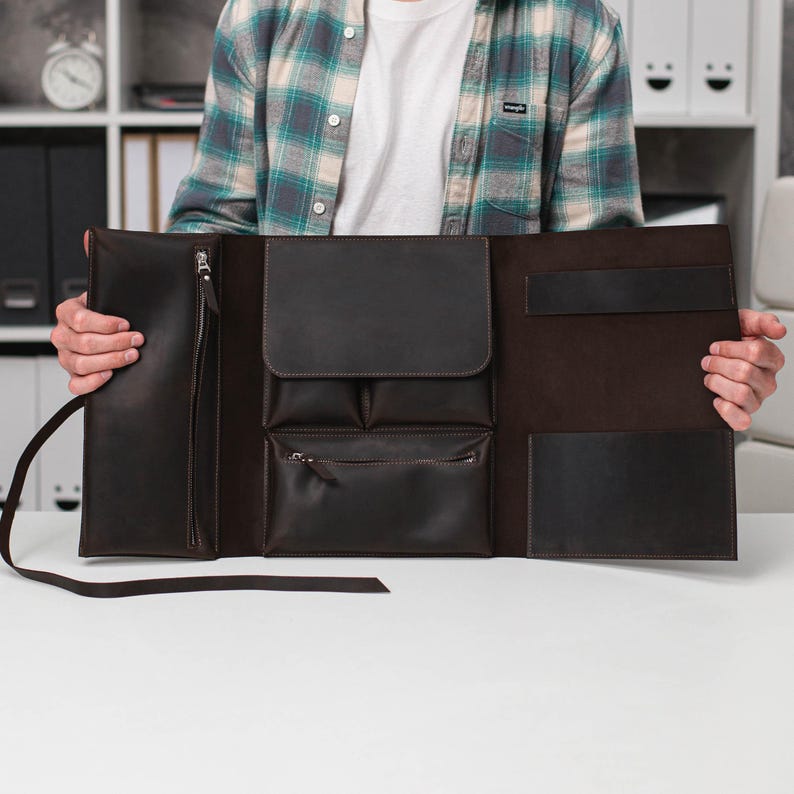 May include: A dark brown leather portfolio with multiple pockets and a zipper closure. The open portfolio displays a variety of compartments, including a large pocket, smaller pockets, and a pen holder. The leather has a smooth texture and a rich, dark color.
