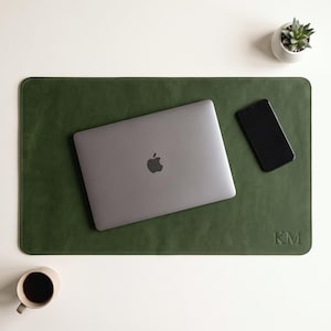 May include: A rectangular olive green leather desk mat with rounded corners and the embossed initials KM in the bottom right corner. A closed grey laptop and a black smartphone are placed on the mat, with a small succulent and a mug nearby.