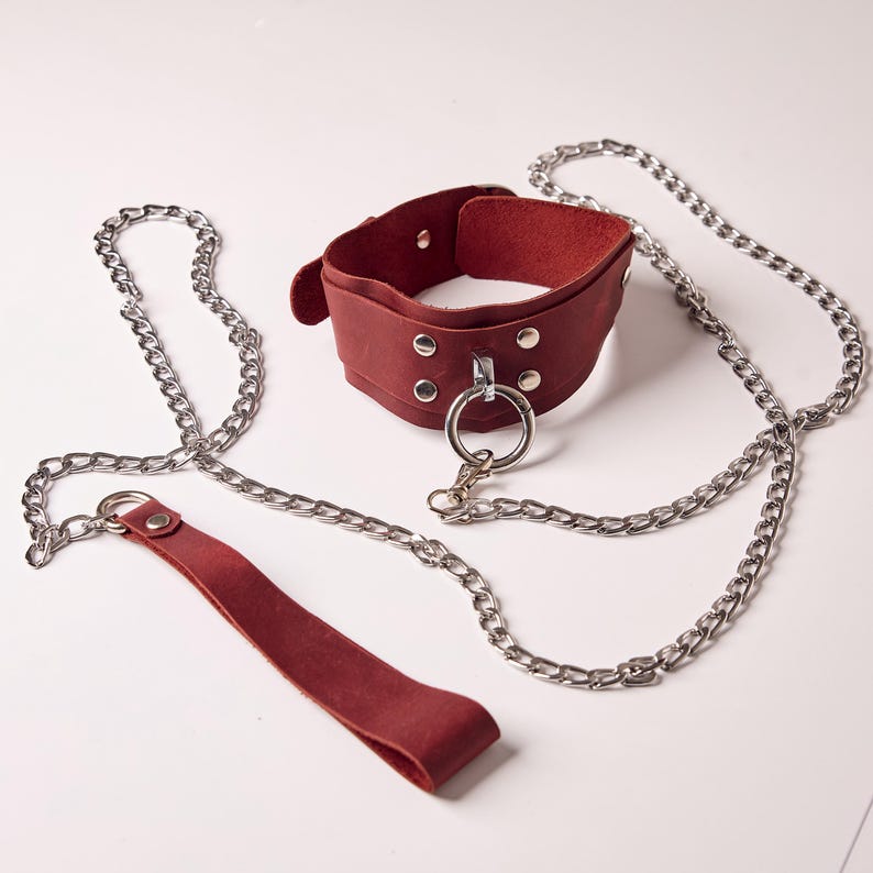May include: A burgundy leather collar with a silver metal ring and chain leash. The collar has a wide band with rivets and a buckle closure. The chain is made of silver metal links, and the leash is made of matching burgundy leather.