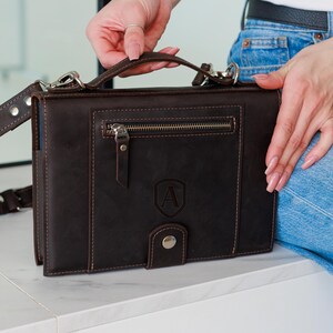 May include: A dark brown leather satchel with a top handle and a detachable shoulder strap. The satchel features a front zipper pocket, a flap closure with a snap, and an embossed letter "A" on the front. The bag has silver-tone hardware and visible stitching.