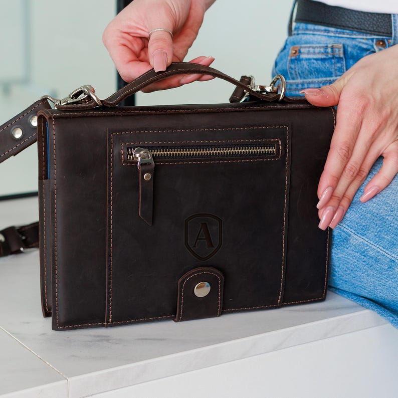 May include: A dark brown leather satchel with a top handle and a detachable shoulder strap. The satchel features a front zipper pocket, a flap closure with a snap, and an embossed letter "A" on the front. The bag has silver-tone hardware and visible stitching.
