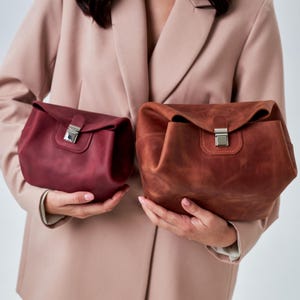 Puede incluir: Dos bolsos de cuero con acabado mate, uno en color burdeos intenso y otro en marrón cálido. Cada bolso cuenta con una solapa plegable asegurada por un cierre de presión de metal plateado, con costuras visibles a lo largo de la lengüeta.