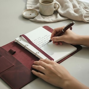 May include: A burgundy leather journal with a white notepad and silver rings. A burgundy ribbon is attached to the notepad. A person is writing with a burgundy pen. A cup of coffee and a small dish are in the background.