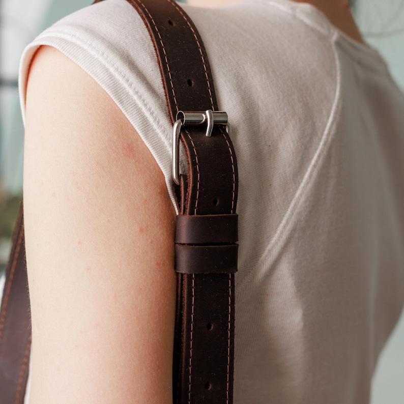 May include: Close-up of a dark brown leather strap with a silver buckle. The strap is stitched with light brown thread. The strap is attached to a white shirt.
