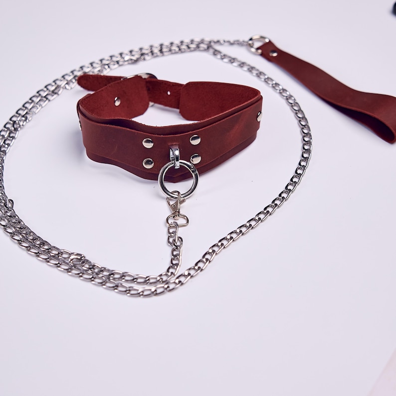 May include: A brown leather collar with silver-toned metal hardware, including a large ring and chain. A matching leather leash is also included. The collar is designed with a curved shape and is secured with a buckle.