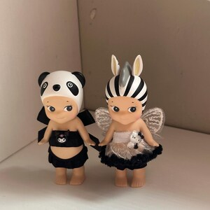 Pu&ograve; includere: Due piccole statuette, una vestita da panda con un completo bianco e nero e l'altra da zebra con un copricapo a righe bianche e nere e ali. Entrambe hanno piccoli fiocchi neri e sono fianco a fianco.