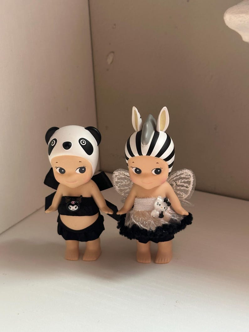 Pu&ograve; includere: Due piccole statuette, una vestita da panda con un completo bianco e nero e l'altra da zebra con un copricapo a righe bianche e nere e ali. Entrambe hanno piccoli fiocchi neri e sono fianco a fianco.