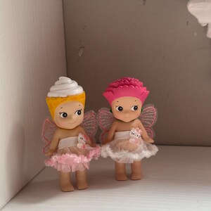 Pu&ograve; includere: Due piccole statuette con grandi occhi e piccole ali. Una ha un cappello a cono gelato giallo e bianco, l'altra un cappello a fiore rosa. Entrambe indossano gonne a balze rosa e bianche e hanno piccole figure di gatto bianco in vita.