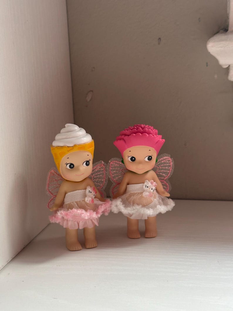 Pu&ograve; includere: Due piccole statuette con grandi occhi e piccole ali. Una ha un cappello a cono gelato giallo e bianco, l'altra un cappello a fiore rosa. Entrambe indossano gonne a balze rosa e bianche e hanno piccole figure di gatto bianco in vita.