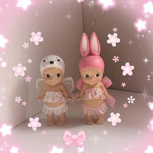 Pu&ograve; includere: Due piccole statuette, una con cappello da foca e ali, l'altra con orecchie da coniglio e un fiocco. Entrambe indossano abiti rosa e bianchi. Lo sfondo &egrave; rosa con decorazioni di stelle e fiori.