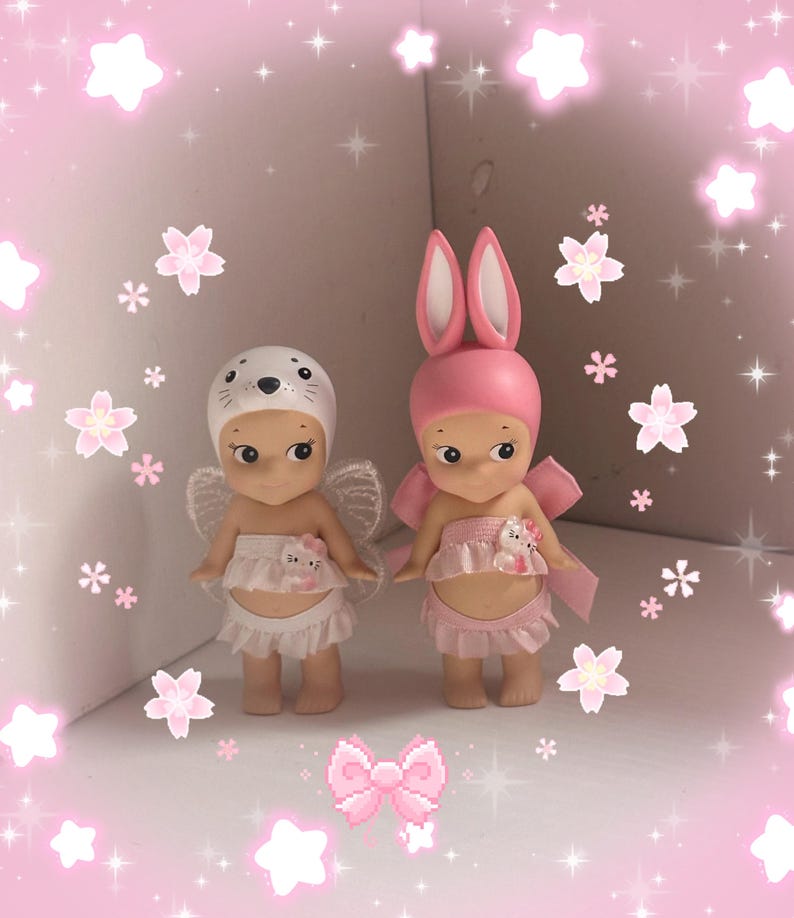 Pu&ograve; includere: Due piccole statuette, una con cappello da foca e ali, l'altra con orecchie da coniglio e un fiocco. Entrambe indossano abiti rosa e bianchi. Lo sfondo &egrave; rosa con decorazioni di stelle e fiori.