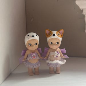 Pu&ograve; includere: Due piccole statuette, ognuna con un cappello a tema animale. Una indossa un cappello da foca, l'altra un cappello da cane. Entrambe hanno ali color lavanda e abiti abbinati. Le statuette sono affiancate.