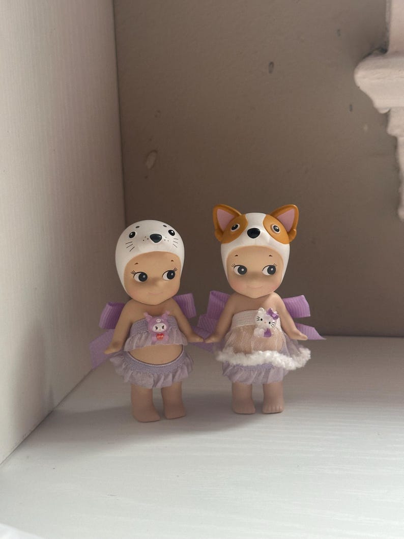 Pu&ograve; includere: Due piccole statuette, ognuna con un cappello a tema animale. Una indossa un cappello da foca, l'altra un cappello da cane. Entrambe hanno ali color lavanda e abiti abbinati. Le statuette sono affiancate.