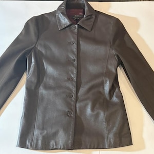 May include: A dark brown leather jacket featuring a classic collar, long sleeves, and a front button closure with five visible buttons. The interior lining is a deep burgundy color, and a black label is visible at the inner neckline.