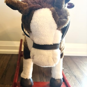 May include: A plush rocking horse with brown and white fur, featuring a white blaze on its face, dark brown hooves, and a black faux leather bridle. The toy is mounted on a red wooden rocker base with two parallel rails and curved supports.