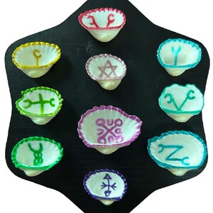 Puede incluir: Una colección de cuencos de conchas marinas blancas con símbolos y bordes bordados de colores, dispuestos sobre una placa de madera negra en forma de hexágono. Los símbolos son de varios colores, incluyendo rojo, amarillo, verde, rosa, azul y morado.