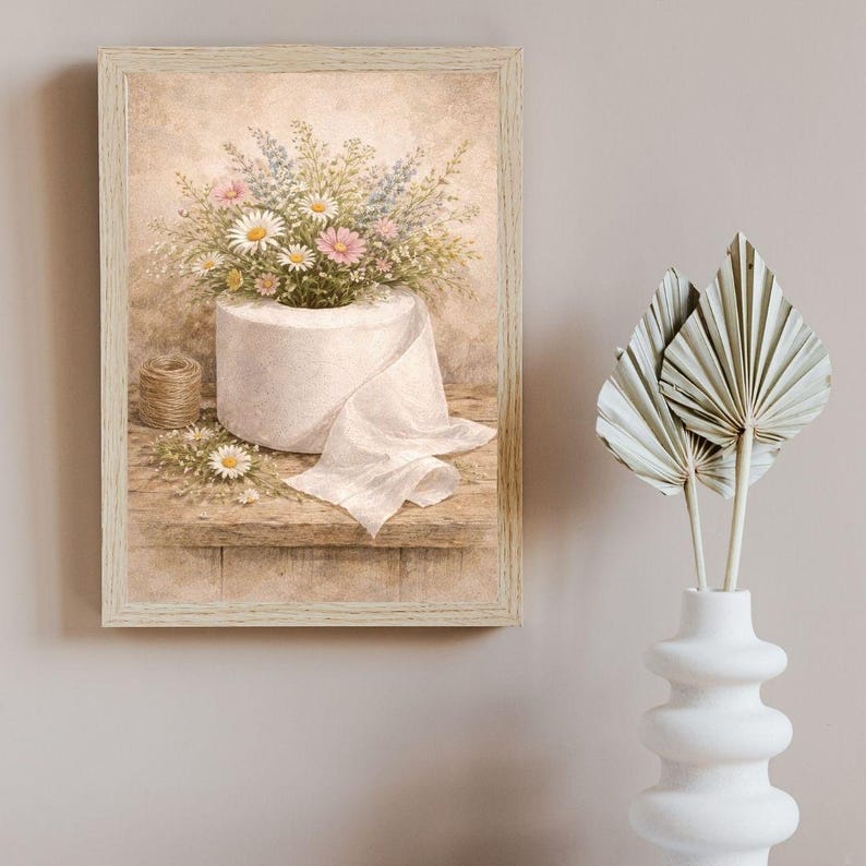 Puede incluir: Una impresi&oacute;n en acuarela enmarcada representa un rollo de papel higi&eacute;nico rebosante de margaritas, flores silvestres rosas y azules. Una peque&ntilde;a bobina de hilo y algunas flores sueltas est&aacute;n sobre una estanter&iacute;a de madera. La obra de arte tiene una est&eacute;tica vintage.
