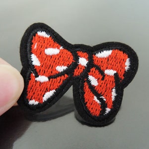 May include: An embroidered patch in the shape of a bow, featuring a vibrant red fabric base with white polka dots and a thick, solid black stitched border outlining the entire shape of the bow design.