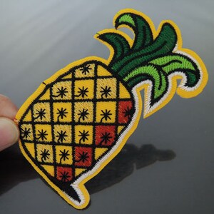 Puede incluir: Parche bordado de piña con cuerpo amarillo, hojas verdes y amarillas y contorno negro. La piña tiene un patrón de cuadrícula con detalles rojos y negros y un borde amarillo.