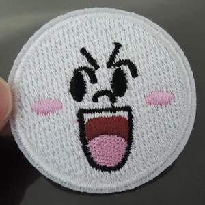 Może przedstawiać: Biała haftowana łatka z kreskówkową twarzą ducha. Duch ma czarne oczy, otwarte czerwone usta i różowe policzki.