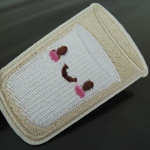 以下が含まれることがあります： 笑顔の顔をした牛乳の入ったコップの白い刺繍パッチ。コップはベージュで縁取られており、顔は茶色の目とピンクの頬をしています。