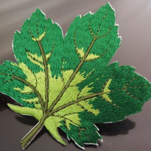 Puede incluir: Parche de hoja bordada en verde y amarillo. La hoja está detallada con venas y un tallo.