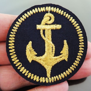 Könnte beinhalten: Ein gold gestickter Anker auf einem schwarzen Stoffpatch mit einem gold gestickten Rand.