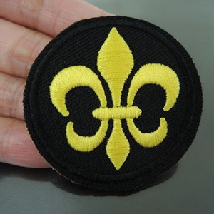Op de afbeelding: Een zwarte strijk-op patch met een geborduurd fleur-de-lis-ontwerp in goud.