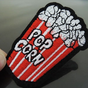 Op de afbeelding: Rode en witte geborduurde strijkpatch met de woorden "POP CORN" en een afbeelding van popcorn in een gestreepte container.