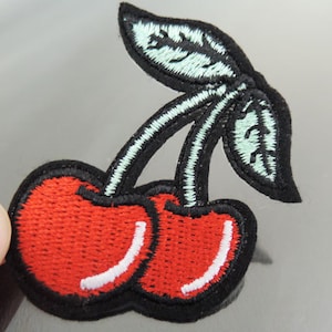 Op de afbeelding: Rode geborduurde kers-patch met groene bladeren en zwarte contouren.