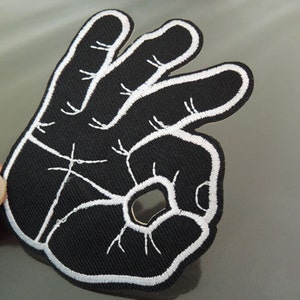 Könnte beinhalten: Schwarzer Stoff-Patch mit weißer Stickerei einer Hand, die das OK-Zeichen macht.
