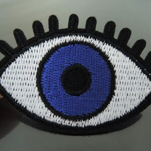 May include: Embroidered blue evil eye patch with white eyelashes and a black pupil. The patch is made of fabric and has a stitched edge.