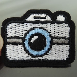 Puede incluir: Parche bordado con forma de cámara. El parche presenta un borde negro, un cuerpo blanco con costuras detalladas, una lente azul y detalles grises. El diseño de la cámara es una representación sencilla y estilizada.