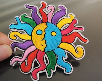 Op de afbeelding: Een kleurrijk geborduurd patch met een gestileerde zon met een gezicht. De zon heeft een geel en blauw gezicht met een rode mond en gele ogen. De zonnestralen zijn gemaakt van meerkleurige borduurdraden.