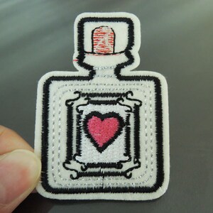 Puede incluir: Parche bordado en fieltro blanco de una botella con una tapa roja y un corazón rosa en su interior. La botella está bordeada con hilo negro y plateado.