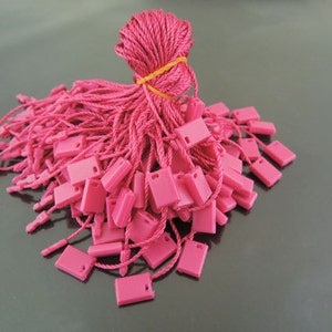 May include: A large pile of pink string tags with a small square plastic tag at the end of each string. The strings are bundled together with a rubber band.