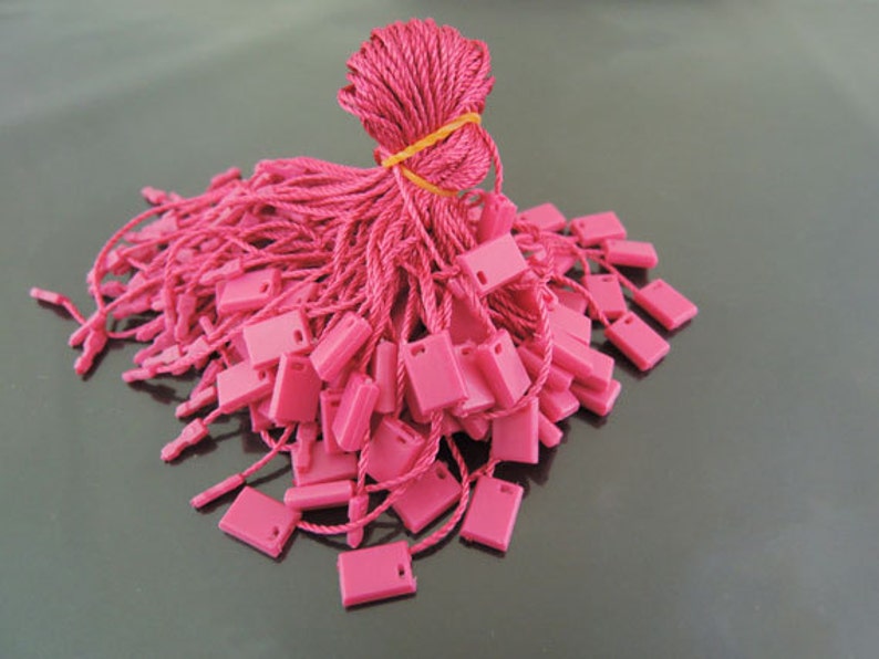 May include: A large pile of pink string tags with a small square plastic tag at the end of each string. The strings are bundled together with a rubber band.