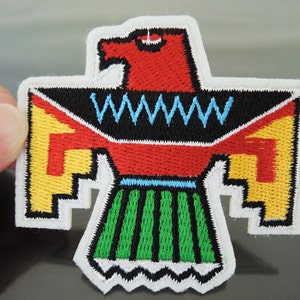 Può includere: Toppa ricamata con un design stilizzato di Thunderbird. La toppa ha una testa rossa, un corpo nero, ali gialle e rosse e una gonna verde e nera. La toppa è bordata di bianco.