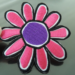 Könnte beinhalten: Gestickter Blumenaufnäher mit einem lila Zentrum, rosa Blütenblättern und schwarzem und weißem Rand. Der Aufnäher wird von einer Hand gehalten, die die leuchtenden Farben und die detaillierten Nähte zeigt. Dieser dekorative Aufnäher eignet sich für Kleidung oder Accessoires.